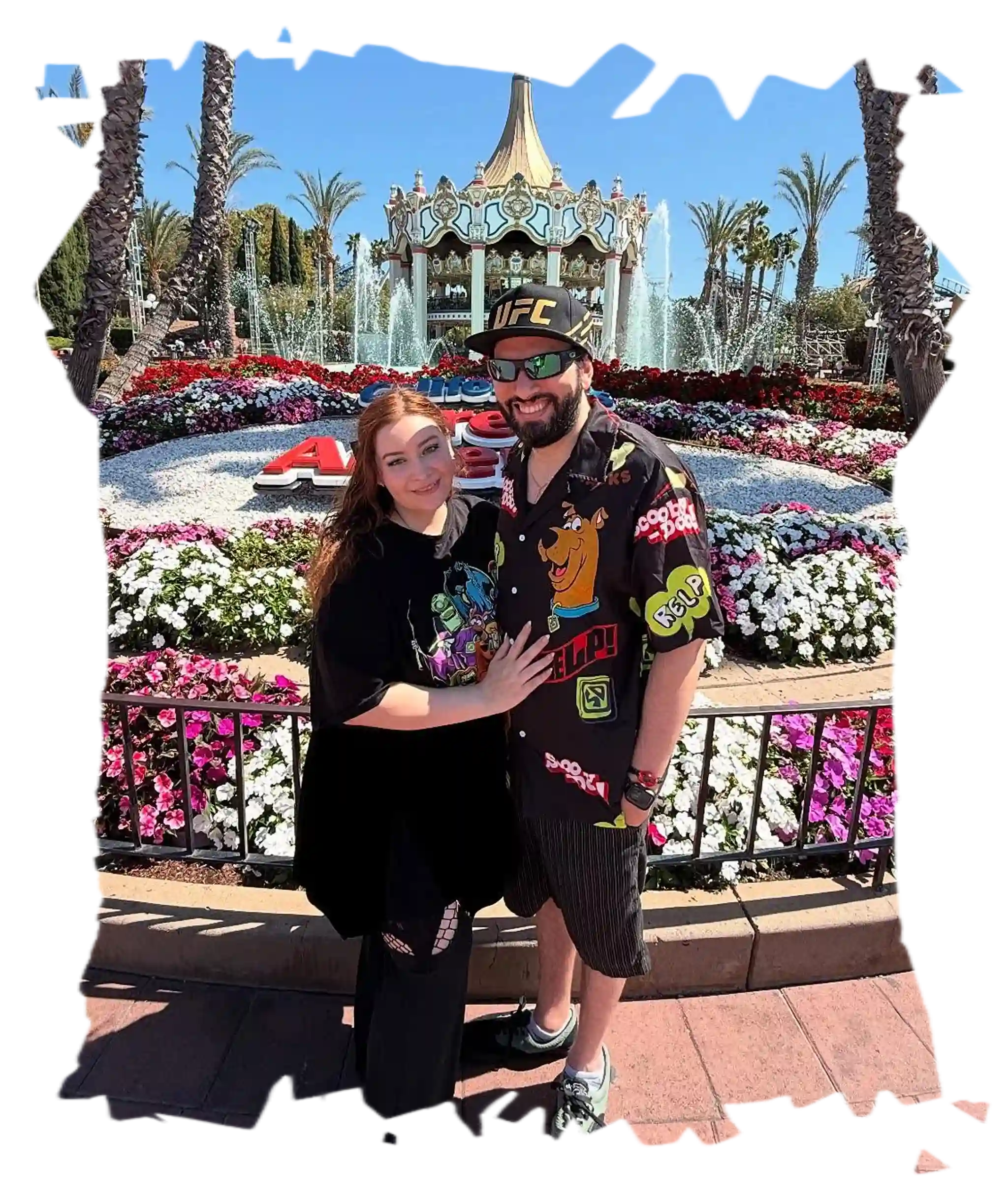 Me and my fiancée Alice smiling together at a theme park in front of a carousel and colorful flowers